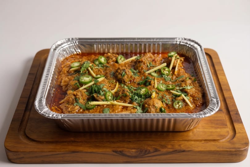 A foil container of chicken karahi, garnished with cilantro, ginger, and green chilies, on a wooden board.