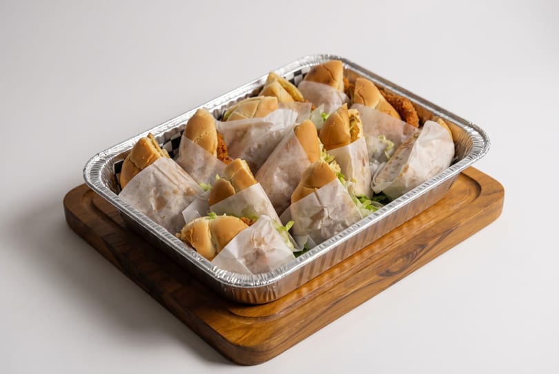 Catering tray filled with individually wrapped chicken sandwiches on a wooden board.