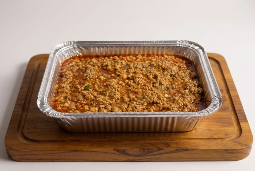 A foil tray filled with a savory ground meat and mushroom mixture, resting on a wooden board.