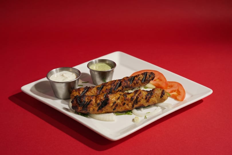 Two grilled seekh kebabs on a white plate with sliced tomatoes, onions, and two dipping sauces.