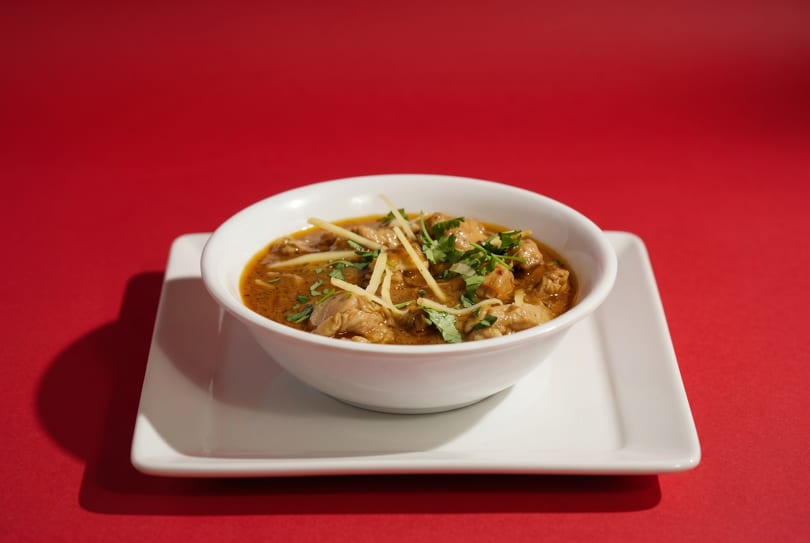 A white bowl of chicken curry with cilantro and ginger on a white plate against a red background.