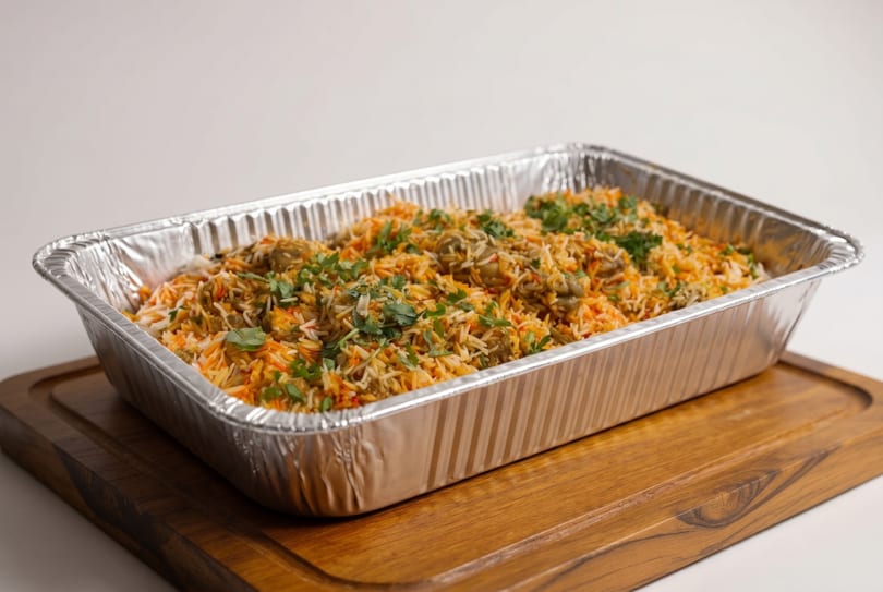 A large aluminum tray filled with chicken biryani, garnished with fresh cilantro, on a wooden board.