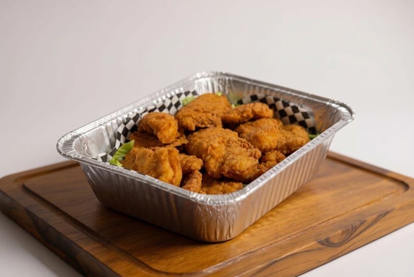 Crispy fried chicken pieces in a foil container with checkered paper, on a wooden board.