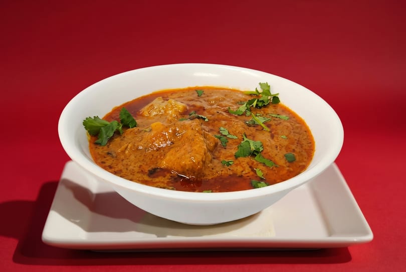 A white bowl of chicken tikka masala, garnished with cilantro, sits on a white plate against a red background.