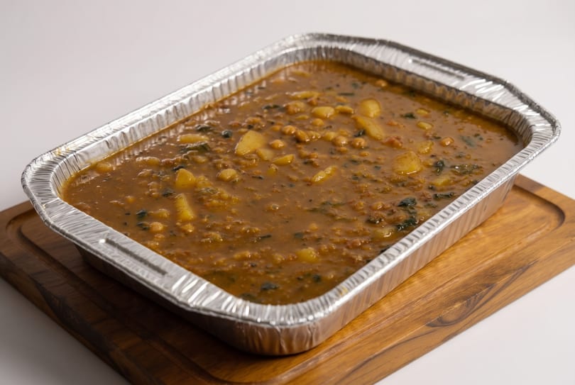 Hearty stew with potatoes and dark greens in a foil pan on a wooden board.