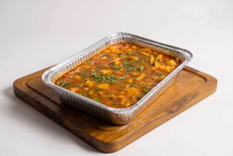 A foil container filled with a hearty potato and vegetable stew, garnished with fresh herbs, on a wooden board.