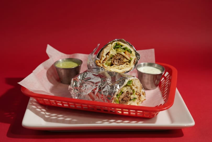 Two foil-wrapped burritos with meat and lettuce, served in a red basket with two dipping sauces.