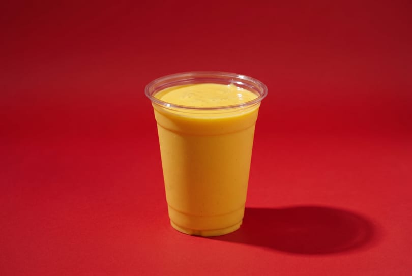 Bright yellow mango smoothie in a clear plastic cup on a vibrant red background.