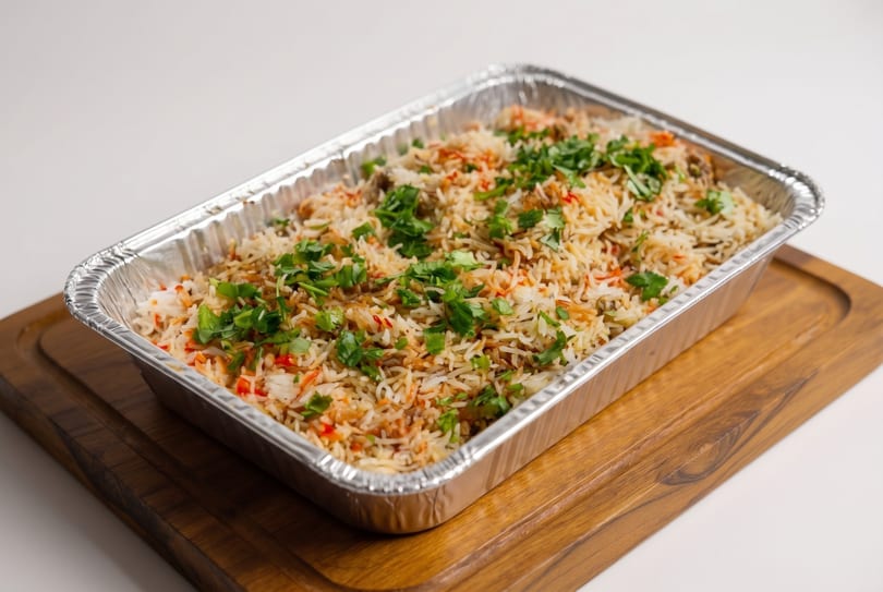 A large aluminum tray of biryani rice with cilantro on a wooden serving board.