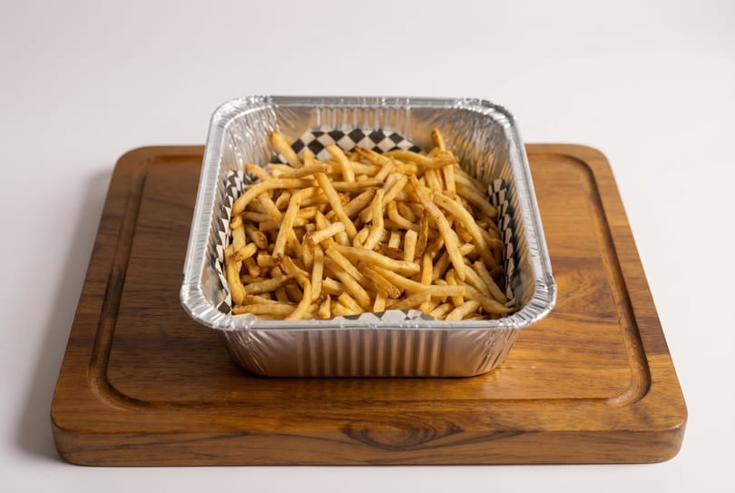 Golden french fries in a foil container with checkered paper, on a wooden cutting board.